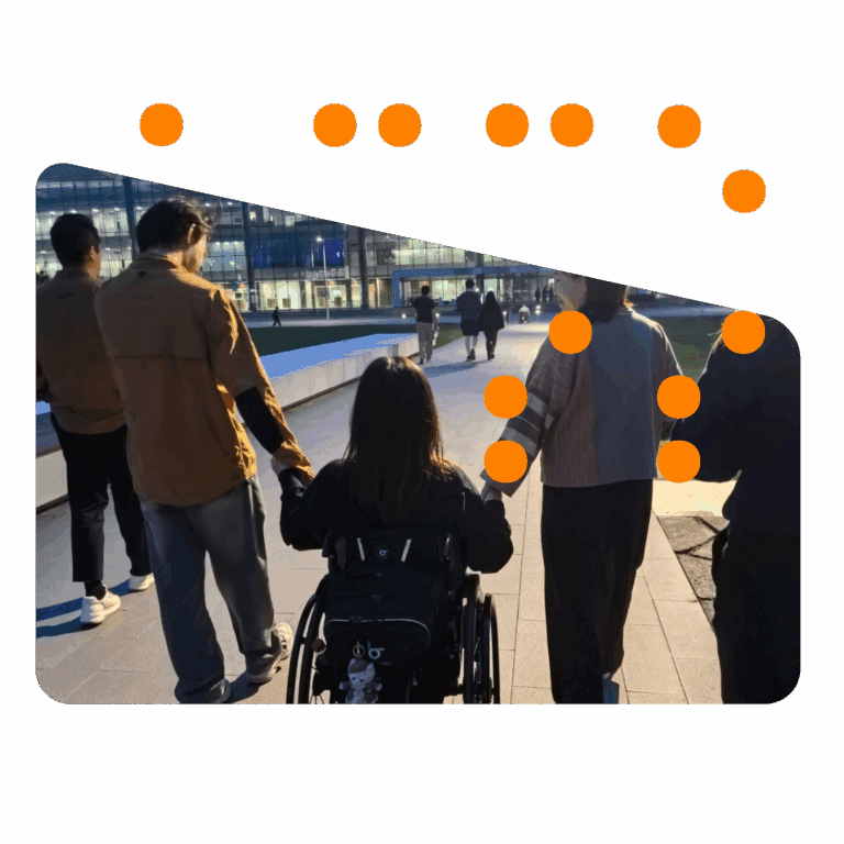 A person using a wheelchair is outdoors in the evening, holding hands with two people walking beside them. They are part of a group moving along a well-lit pathway in front of a modern glass building.