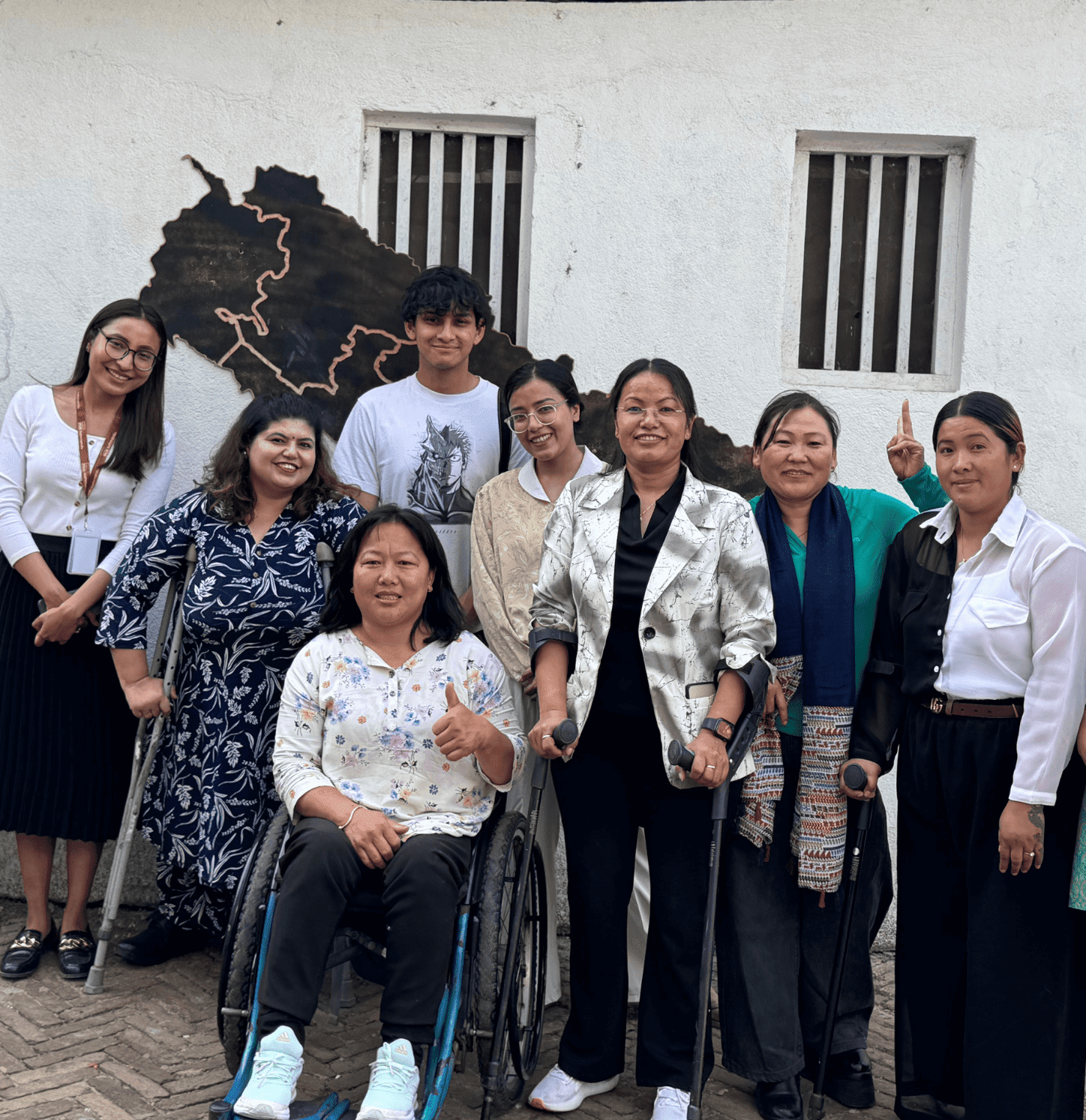 A diverse group of eight people, including two women in wheelchairs, pose together and smile in front of a white wall with a metal map art installation. The group includes both men and women, dressed in a mix of casual and professional attire, suggesting a friendly or inclusive event setting.