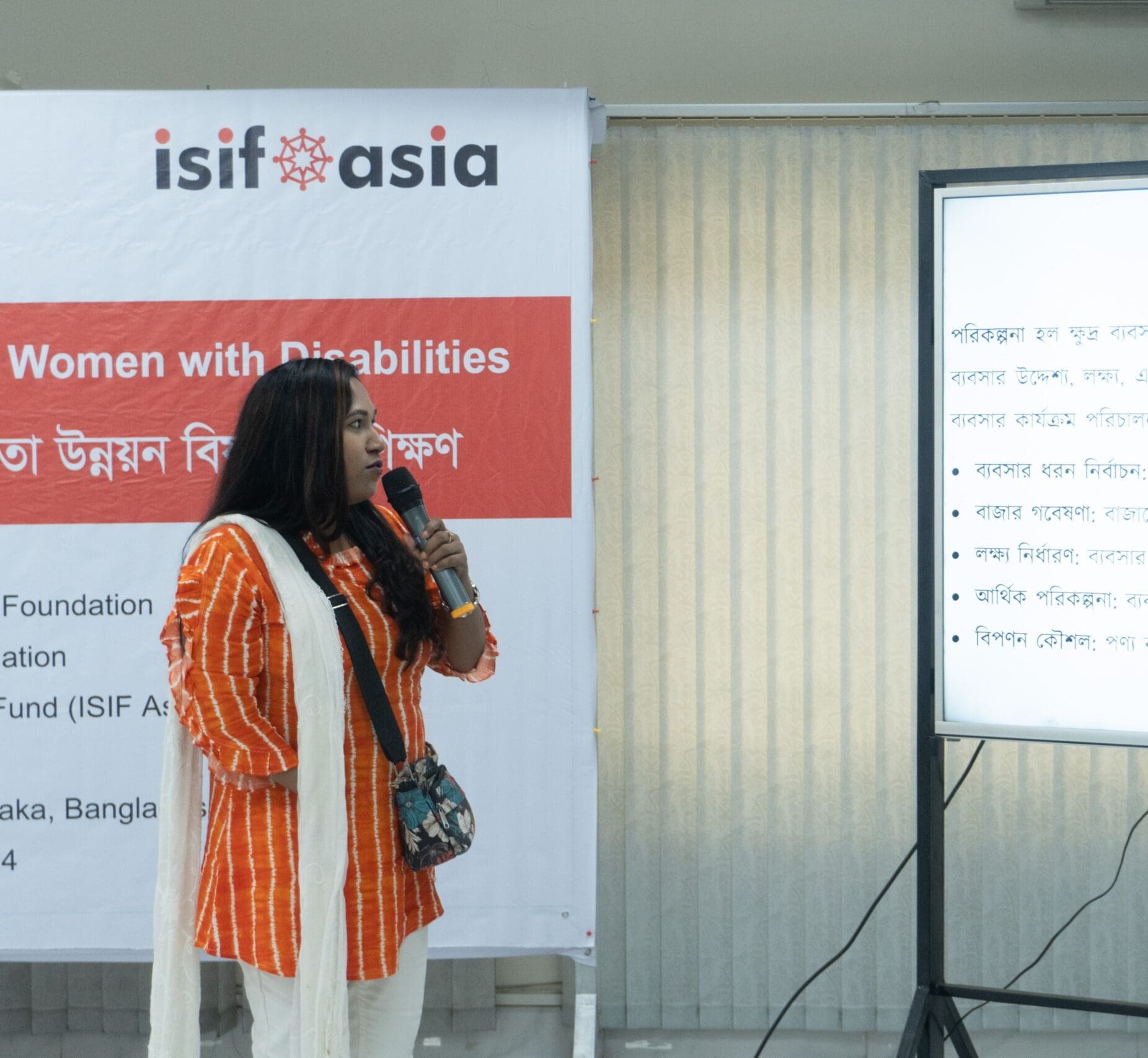A woman with a physical impairment, dressed in an orange and white outfit, speaks into a microphone while presenting at an event. She stands beside a screen displaying text in Bengali.