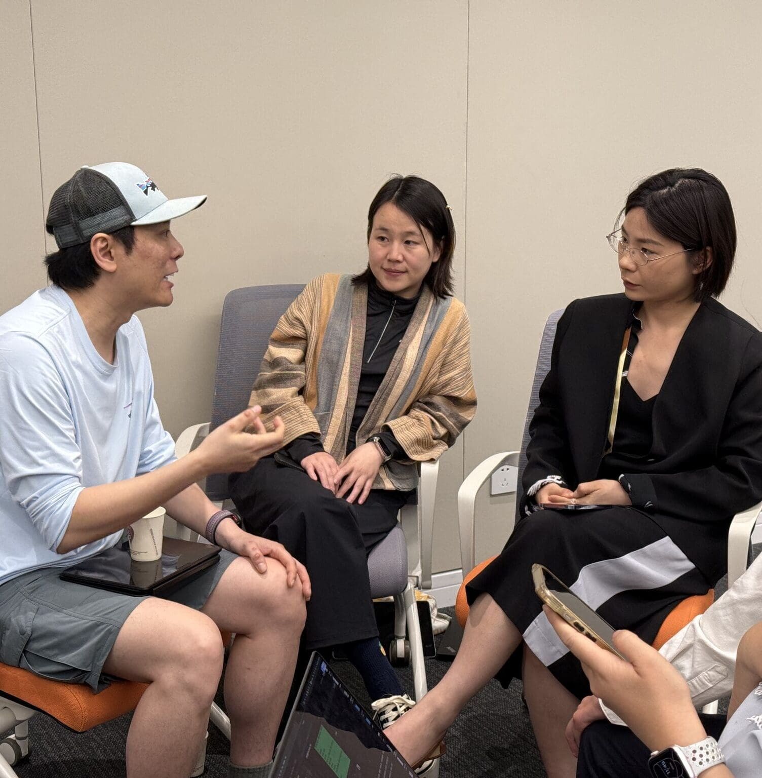Four people sit in a small circle having a focused conversation. One man, casually dressed in a cap and shorts, gestures while speaking. The others—three women—listen attentively, with one holding a phone and another seated with legs crossed. The setting appears to be an informal indoor meeting or workshop.